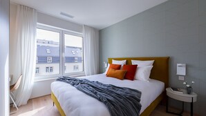 Ein Hotelzimmer mit einem großen Bett, einem Nachttisch mit Lampe und Blick auf Gebäude durch das Fenster.