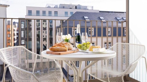 Ein Balkon mit einem Tisch, gedeckt für eine Mahlzeit, mit Brot, Käse, Trauben und einer Flasche.