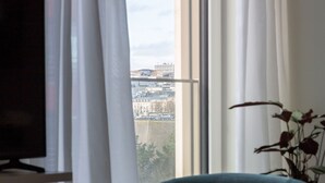 A room with a view of a cityscape through large windows, a television on a stand, a green chair, and a small table with a book. - Deluxe Studio      . (Luxembourg)