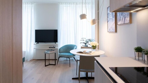 A modern living room with a flat-screen TV, a blue armchair, a round dining table, and a small side table with books. - Deluxe Studio      . (Luxembourg)