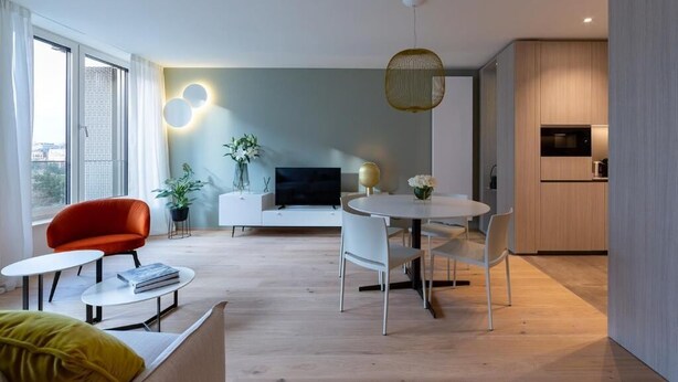 Una sala moderna con un sofá, una butaca roja, una mesa de comedor redonda con sillas, un televisor y una zona de cocina con armarios de madera.