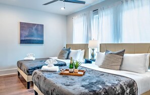Une chambre d’hôtel avec deux lits, une grande fenêtre avec des rideaux, un ventilateur de plafond, une table de chevet avec une bouteille et un plateau, et un tableau au mur.