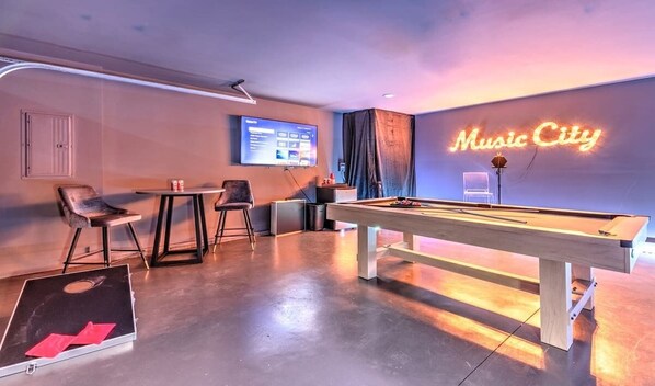A room with a pool table, chairs, a TV screen, and a neon sign reading "Music City".