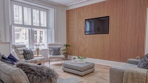 A modern living room with a large window, a flat-screen TV mounted on a wooden wall, and a cozy seating area with a fur throw on the sofa. - Luxury Townhouse in the centre of St Andrews (St Andrews)