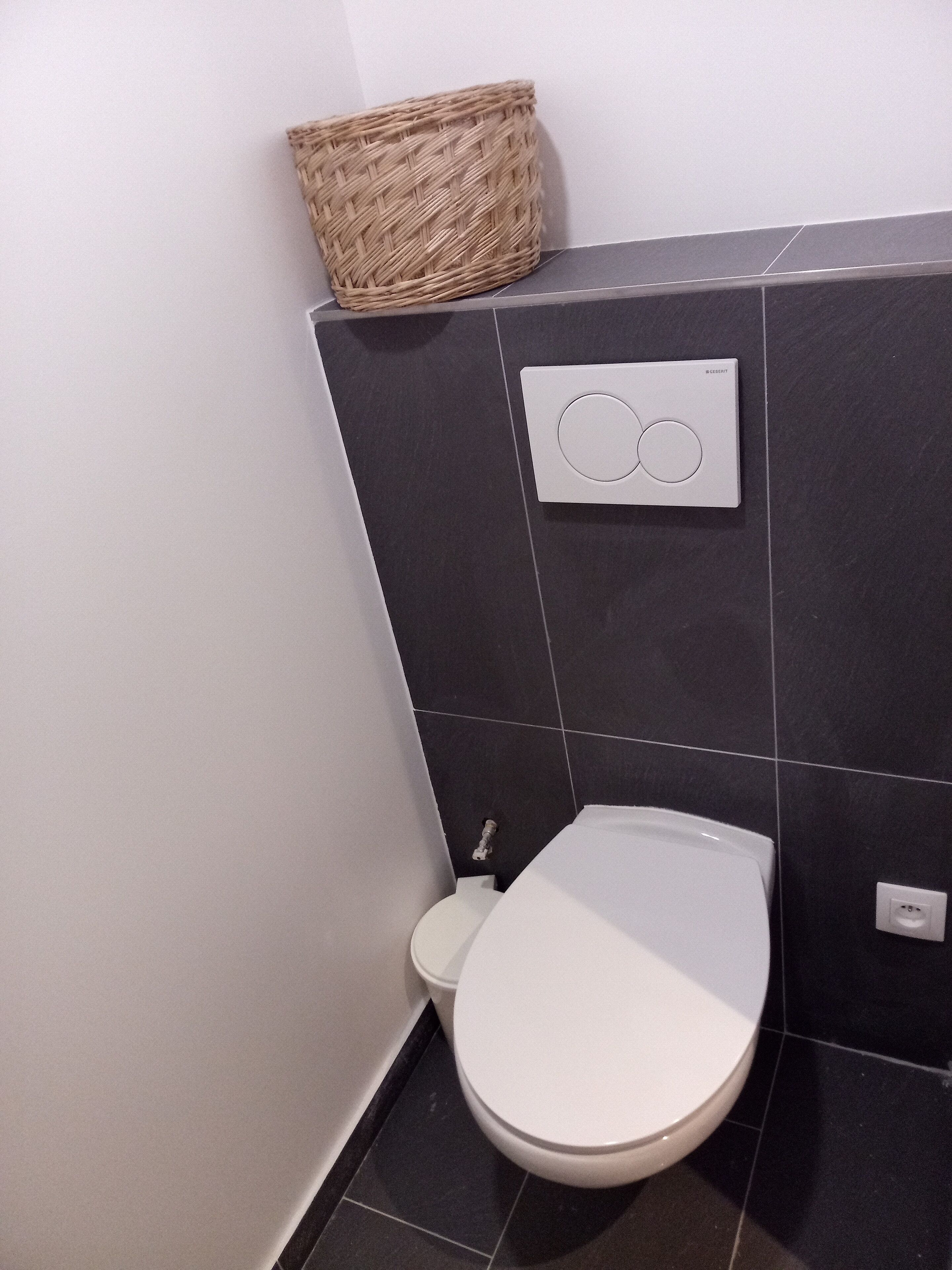 Moderni kylpyhuone, jossa on valkoinen wc-istuin, tummat laattaseinät ja seinällä roikkuva punottu kori.