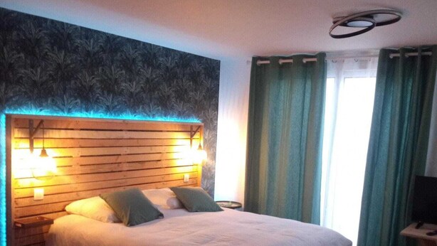 Un dormitorio con cabecera de madera, dos lámparas de noche, una cama con ropa de cama blanca, una mesita de noche pequeña, un televisor sobre un soporte y una ventana con cortinas.