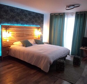 Une chambre à coucher avec une tête de lit en bois, deux lampes de chevet, un lit avec une literie blanche, une petite table de chevet, un téléviseur posé sur un meuble et une fenêtre avec des rideaux.