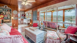 Een gezellig interieur van een chalet met houten wanden, een groot raam met uitzicht op een meer, een plafondventilator, en diverse meubels waaronder een rode bank en een fauteuil met bloemenprint.