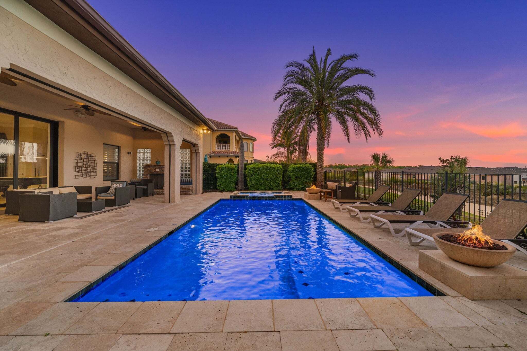 A swimming pool with lounge chairs and a fire feature, surrounded by a house with a patio and palm trees.