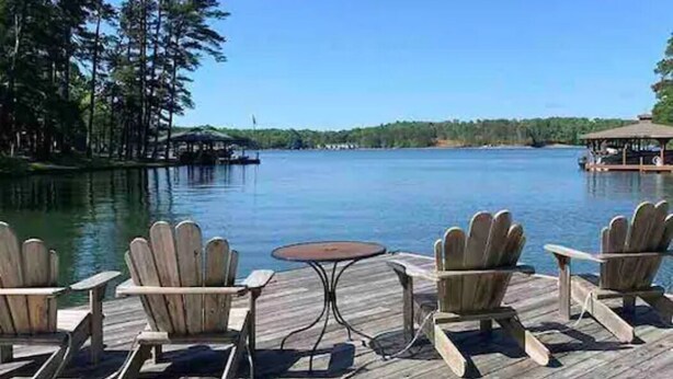 A wooden deck with Adirondack chairs and a small table, overlooking a calm lake with trees and a distant pier. - Castaway Cottage (Eclectic)
