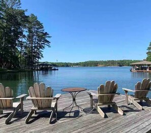 Une terrasse en bois, agrémentée de chaises Adirondack et d’une petite table, offre une vue sur un lac paisible, bordé d’arbres et d’une jetée située au loin.