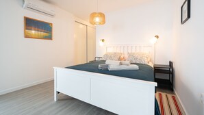 Una camera da letto moderna con un letto bianco, un comodino, un tavolo di fianco al letto, un condizionatore d'aria a muro, una lampada a sospensione e un quadro incorniciato appeso al muro.