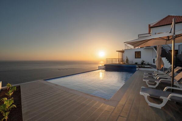 A poolside view with a sunset over the sea, a building with a balcony, and lounge chairs. - Nearer to the Sea than the sky, an amazing views and peaceful escape. (Arco da Calheta)