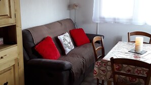 Et hyggeligt værelse med en brun sofa, røde og hvide puder, et træskab og et bord med et mønstret duge.