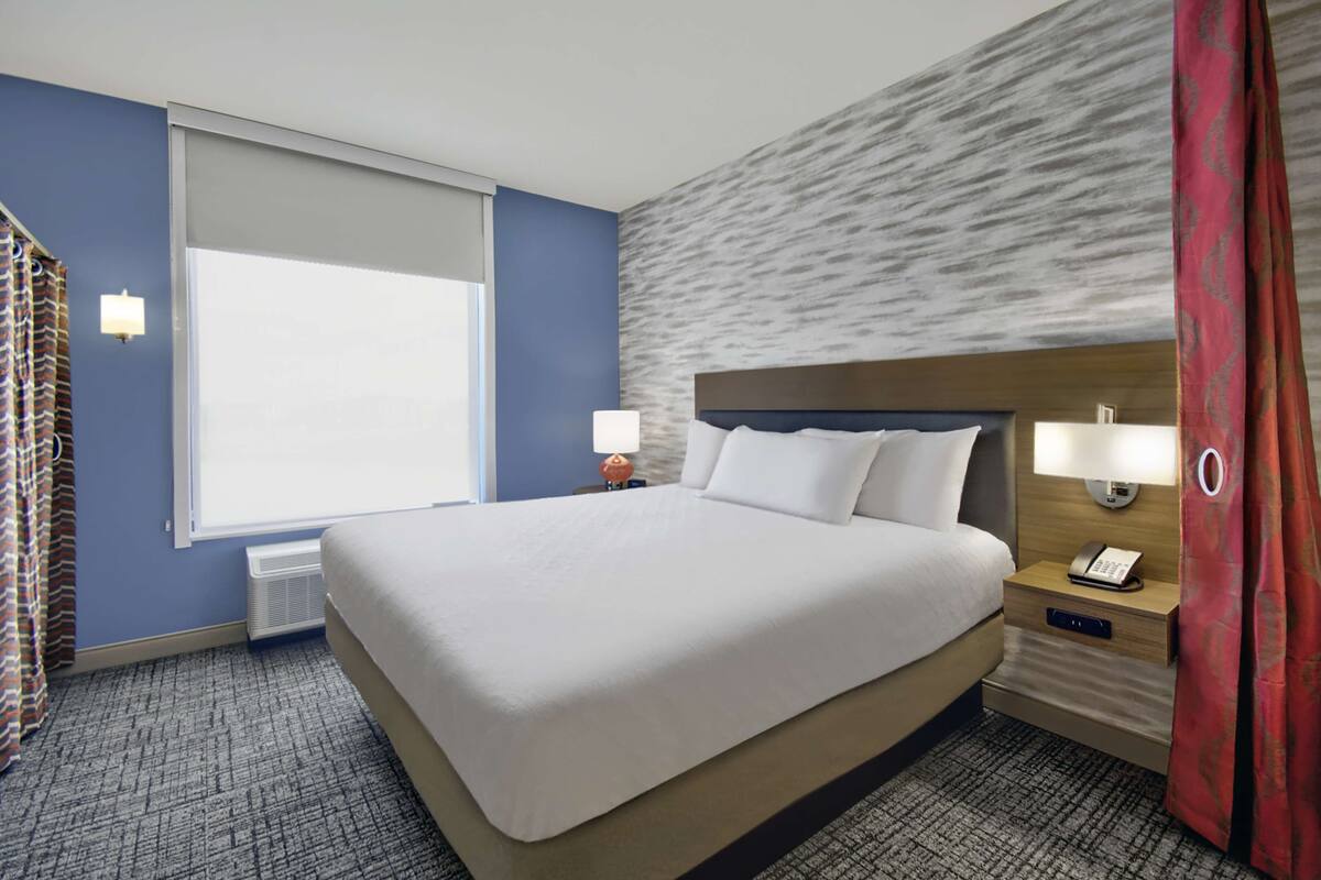 Una habitación de hotel con una cama grande, una mesita de noche con teléfono, una ventana con cortinas y una pared con textura decorativa.