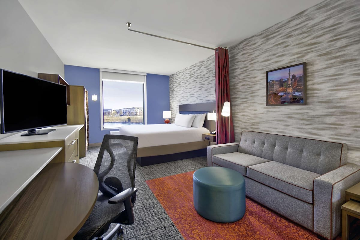 Habitación de hotel con cama, sofá, escritorio con televisor y ventana grande.