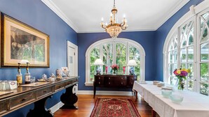 Ein Zimmer mit einer dunkelblauen Wand, einem Chandelier, einem großen Fenster mit weißen Rahmen, einem Holztisch mit weißem Tischlauf und einem gemusterten Teppich.