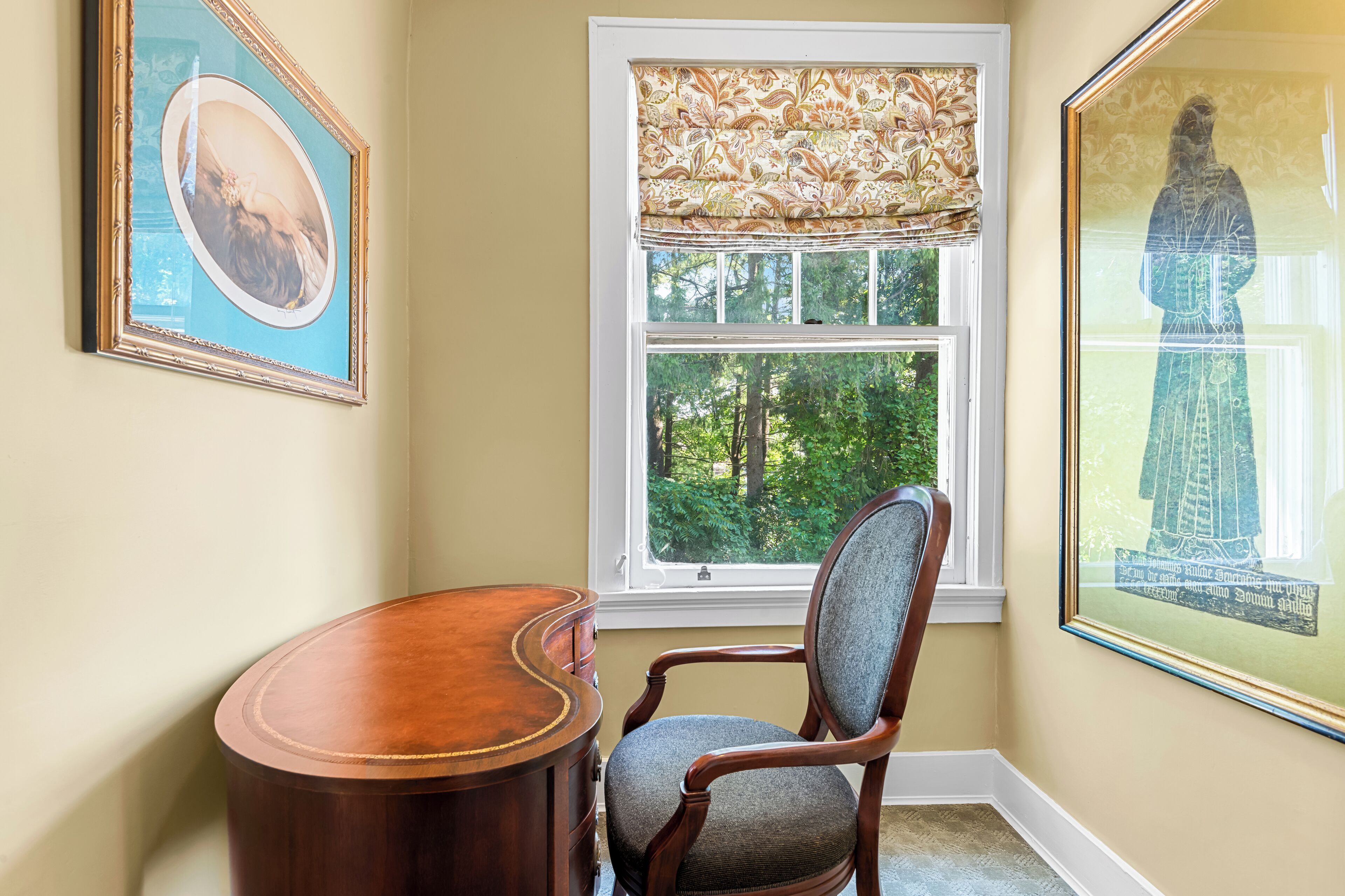 A corner room with a window, a chair, a desk, and two framed artworks.