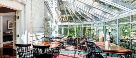 A spacious room with a high ceiling and large windows, featuring a long table and several chairs, surrounded by a patterned carpet.