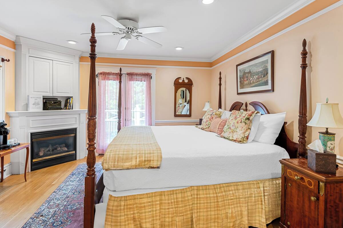 A bedroom with a four-poster bed, a fireplace, a ceiling fan, and a large window with curtains.