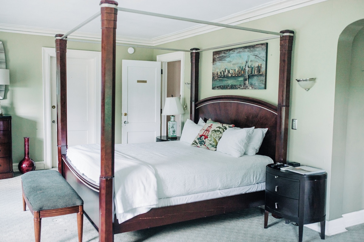 A four-poster bed with a wooden frame and white bedding, a small bedside table with drawers, a bench, and a vase on the floor.
