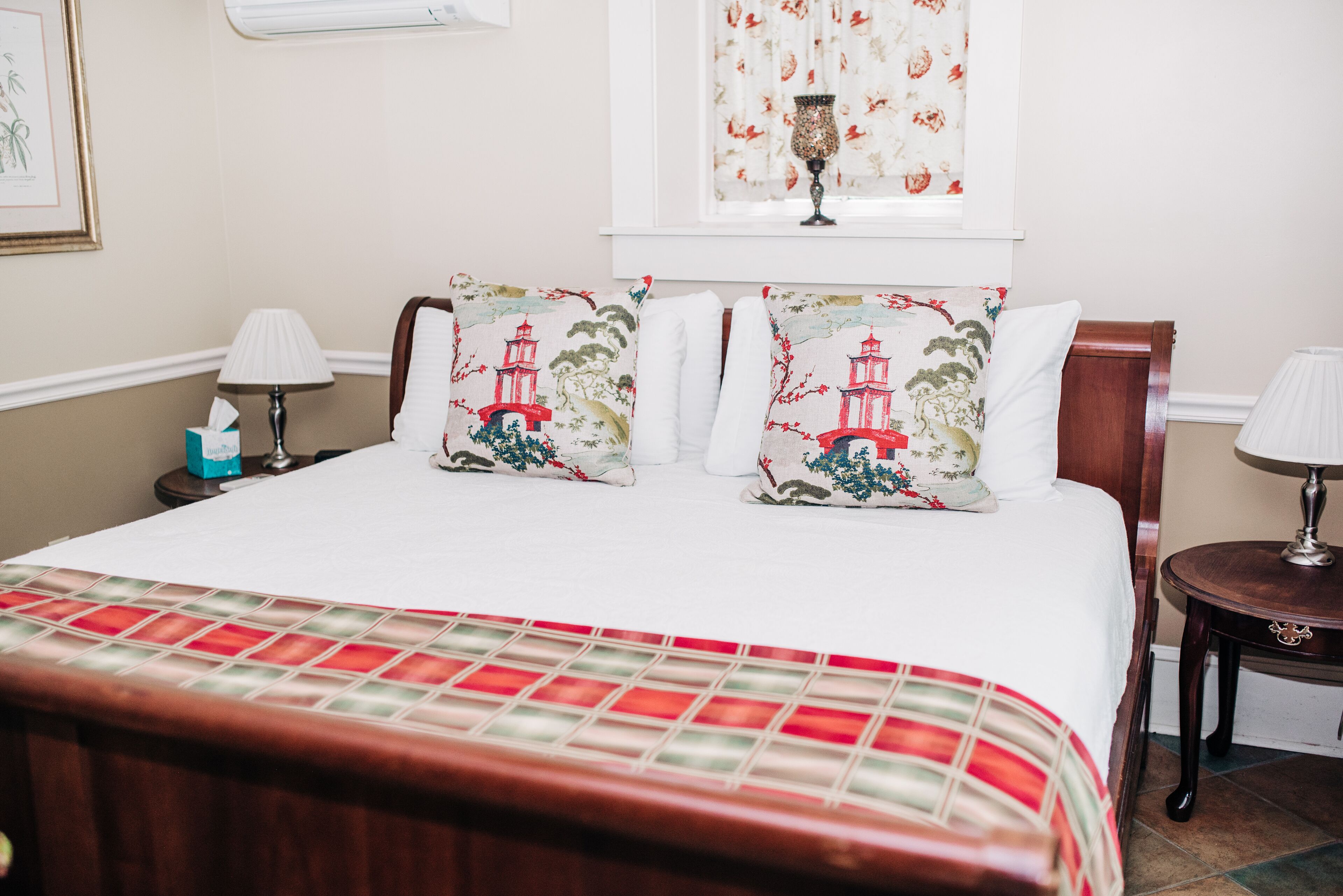 Una cama bien tendida con una colcha a cuadros rojo y blanco, dos almohadas decorativas con diseño de faro, una mesita de noche con lámpara y una ventana con cortinas florales.
