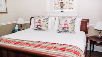 Una cama bien tendida con una colcha a cuadros rojo y blanco, dos almohadas decorativas con diseño de faro, una mesita de noche con lámpara y una ventana con cortinas florales.