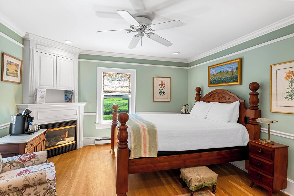 A bedroom with a wooden bed, a nightstand, a dresser, a fireplace, and a window with a view of greenery.