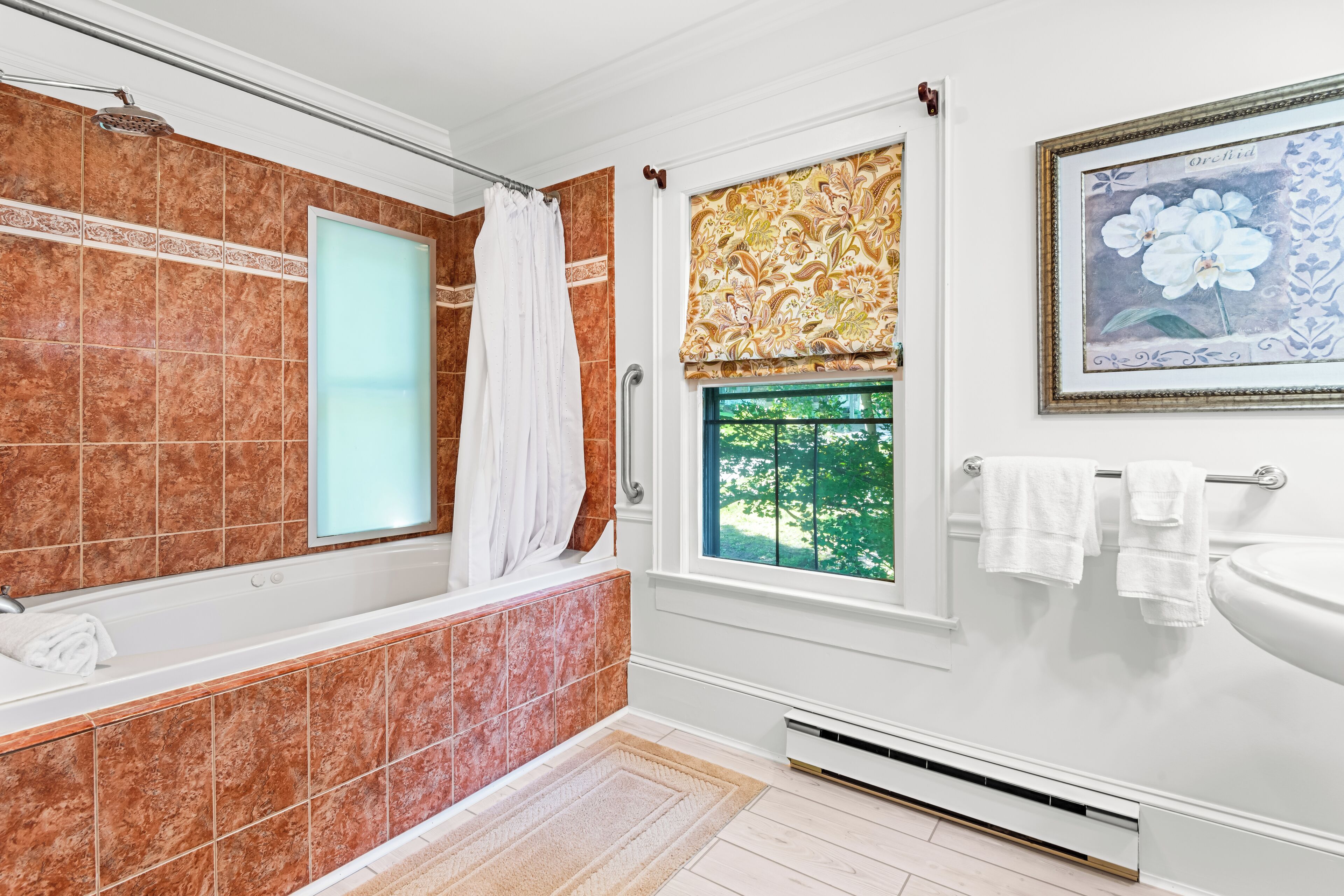 Ein Badezimmer mit Badewanne, einem Fenster mit Blick ins Grüne, einem Waschbecken und einem Bild einer Orchidee im Bilderrahmen.