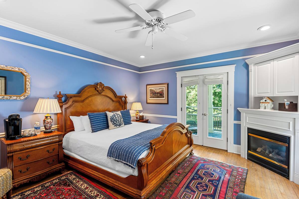 A bedroom with a wooden bed, a ceiling fan, a fireplace, and a view of the outdoors.
