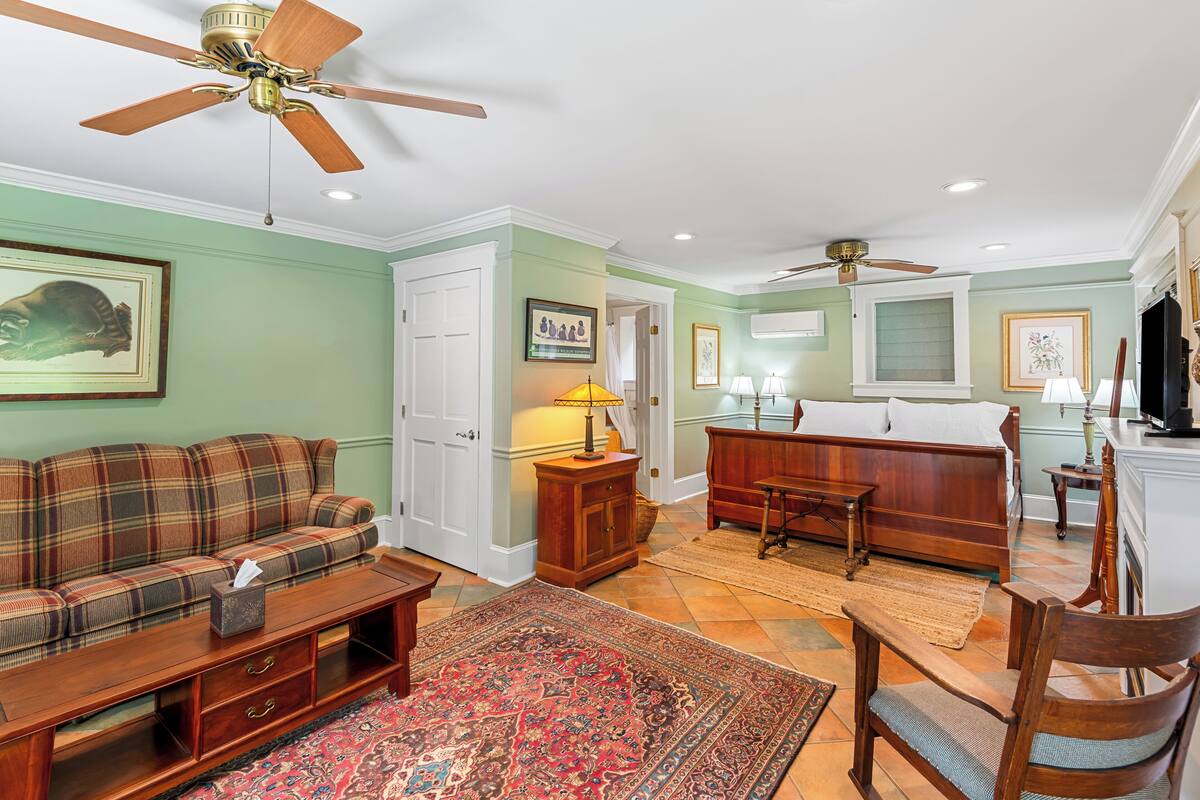 A spacious living room with a sofa, a wooden coffee table, a patterned rug, a bed with a wooden headboard, and a ceiling fan.