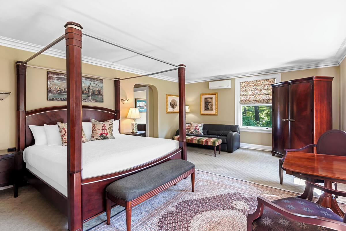A four-poster bed with a bench, a dining table with chairs, a sofa, and a cabinet in a well-lit room with artwork on the walls.