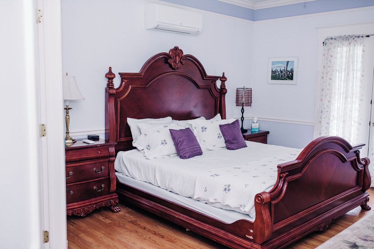 A wooden four-poster bed with a white bedspread and purple pillows, a nightstand with a lamp, and a wall-mounted air conditioner.