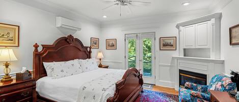A bedroom with a large wooden bed, a ceiling fan, an air conditioning unit, a fireplace, and a view of the outdoors through a door.