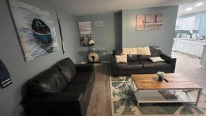 Un salon avec un canapé en cuir noir, une table basse et un tapis. Un tableau représentant un bateau est accroché au mur, et on aperçoit une cuisine au fond de la pièce.