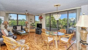A spacious living room with a large window, a ceiling fan, a television, and a dining area with wicker chairs and a glass table. - Anchorage 204 (Siesta Key)
