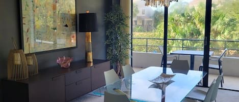 Uma sala de jantar moderna com uma mesa de vidro, cadeiras de metal, um candelabro, um grande quadro abstrato, um abajur e um vaso com planta.
