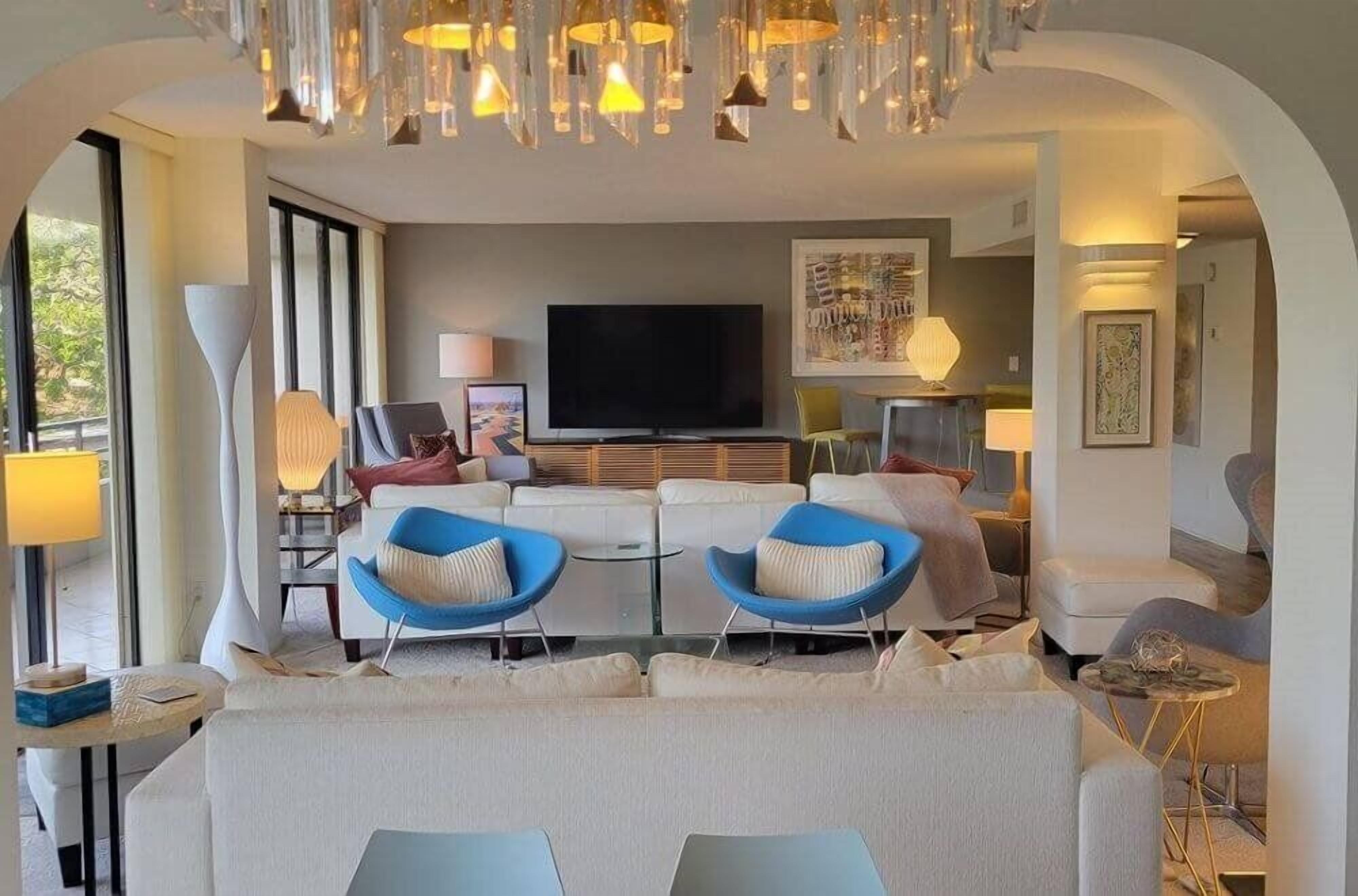 A modern living room with a large TV, two blue armchairs, a sofa, and a chandelier.