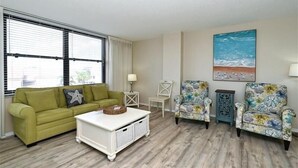 Ein Wohnzimmer mit einem grünen Sofa, einem weißen Couchtisch, zwei floralen Sesseln und einem Gemälde, das eine Strandlandschaft zeigt.