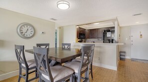 A dining area with a wooden table and grey chairs, a wall clock, and a kitchen with dark wooden cabinets and a stainless steel refrigerator. - Anchorage 405 (Siesta Key)