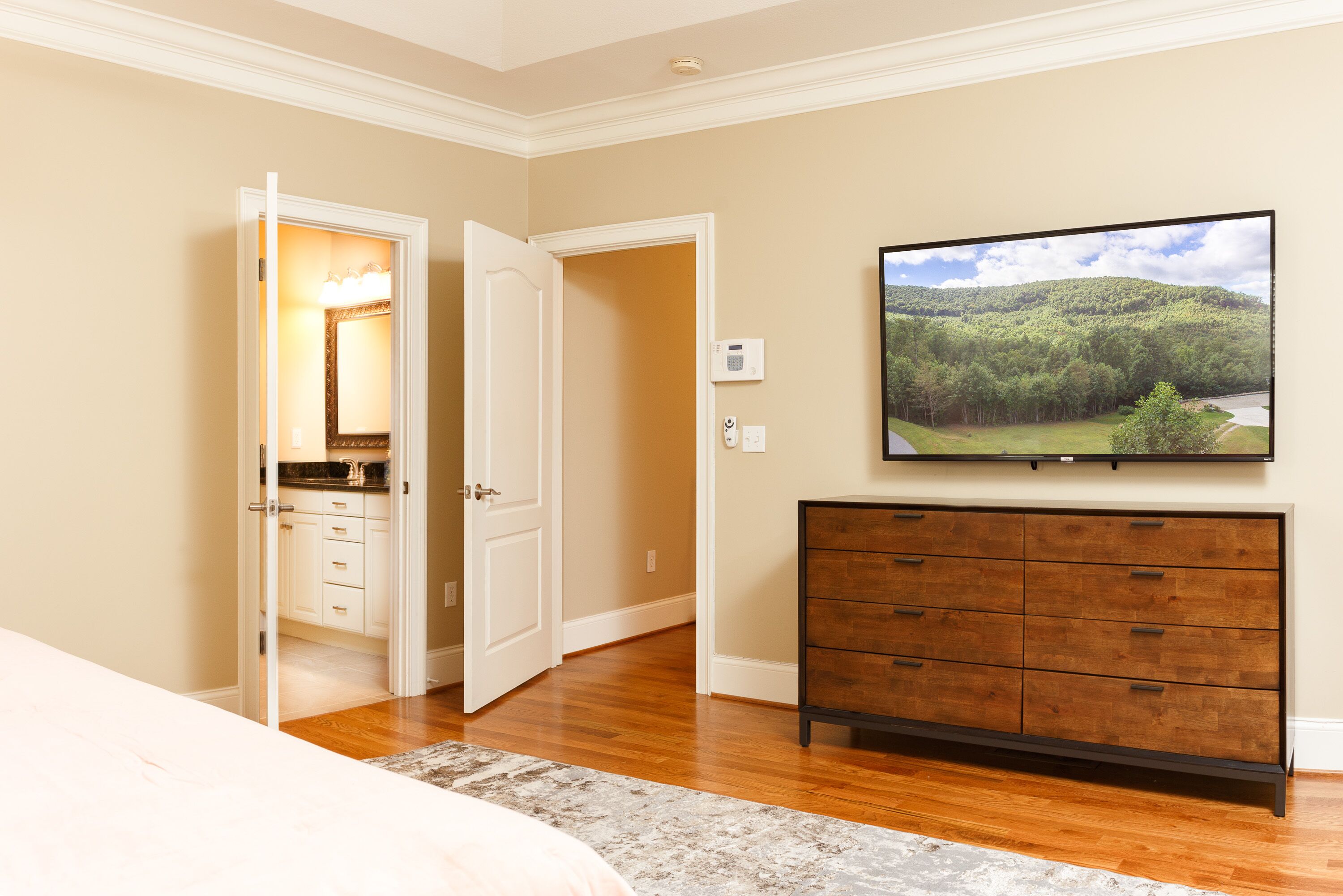Une chambre à coucher avec une commode en bois, un téléviseur fixé au mur et une porte donnant sur une salle de bain.