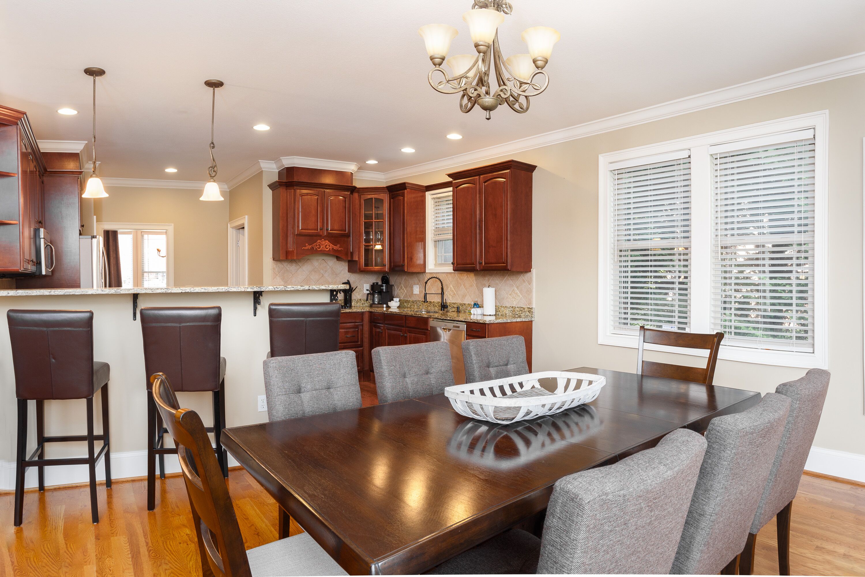 A modern kitchen with dark wooden cabinets, a large island, and a dining area with a wooden table and grey upholstered chairs.