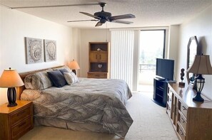 Une chambre à coucher comprenant un lit, deux lampes de chevet, un ventilateur de plafond, une armoire en bois, un téléviseur et des œuvres d’art accrochées au mur.
