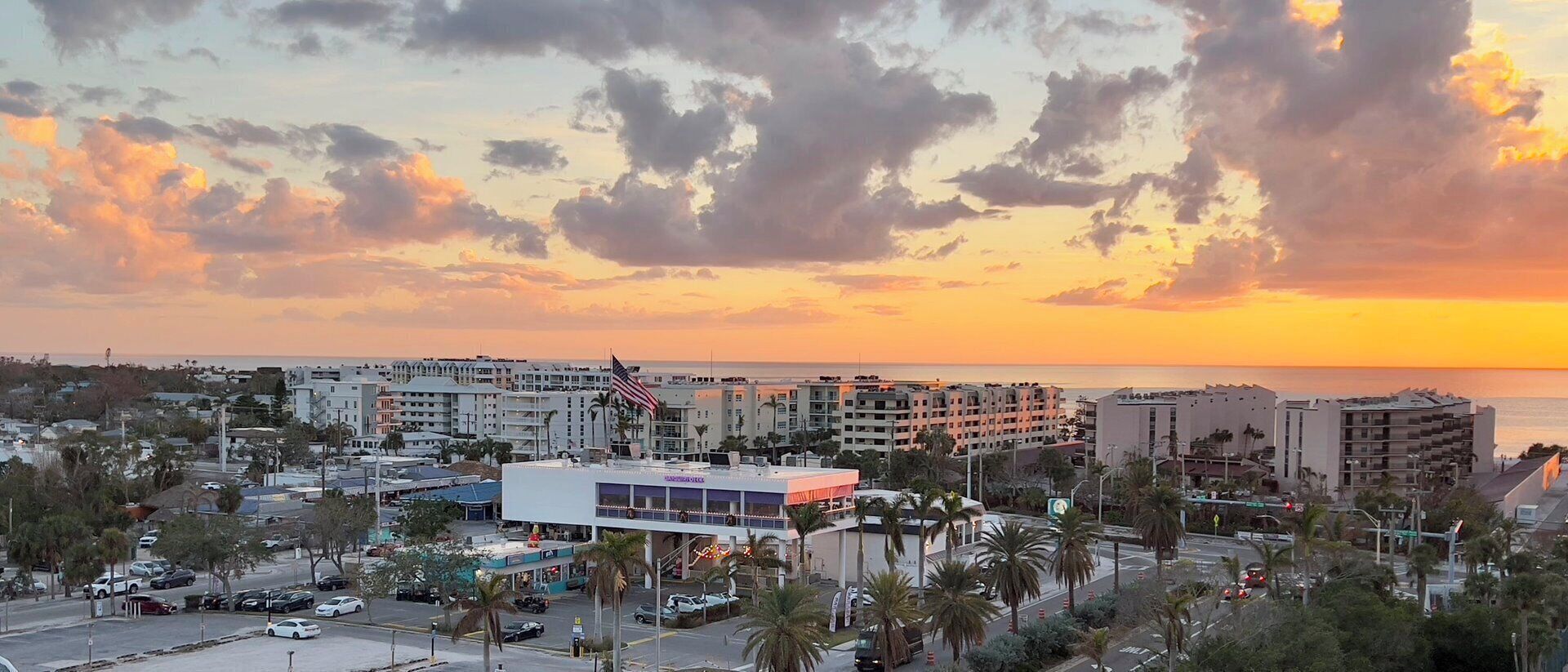 A coastal cityscape with a sunset, featuring multi-story buildings, a prominent hotel with a red roof, and palm trees.