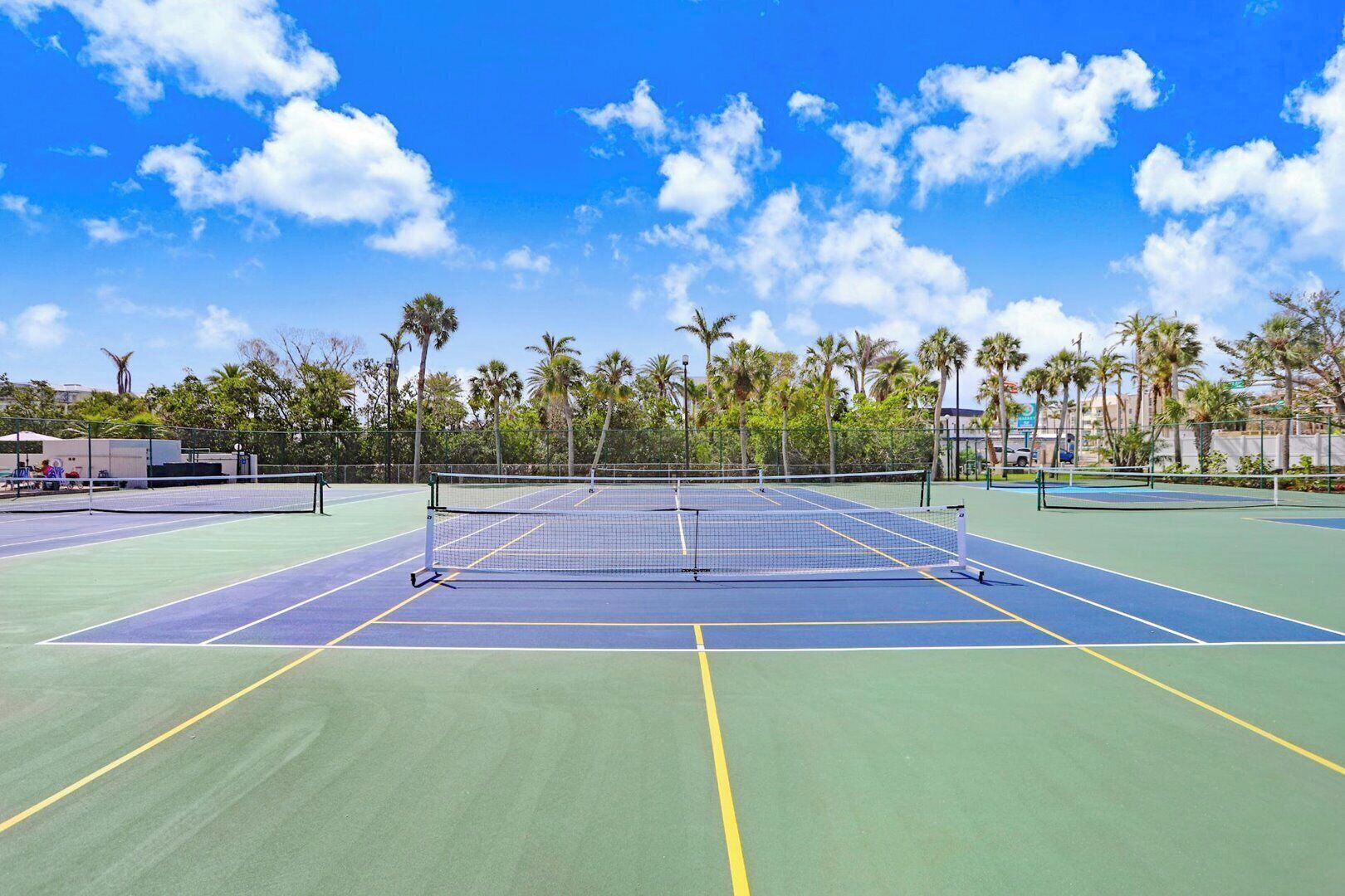 Un court de tennis avec un filet au centre, entouré de palmiers et surplombé d’un ciel bleu azur.