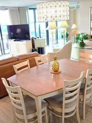 Un salon moderne avec un canapé en cuir marron, une table à manger en bois et des chaises blanches, un centre de table en forme d’ananas et une grande fenêtre avec des rideaux.