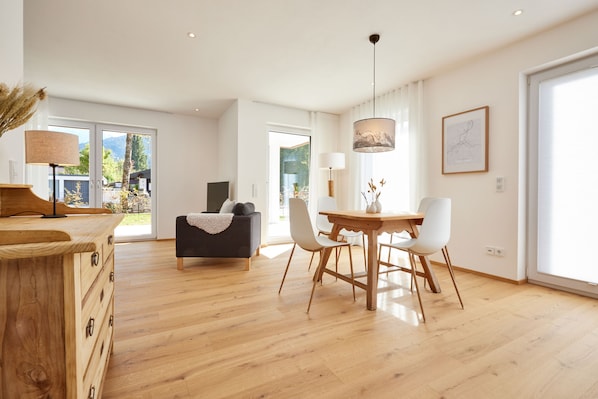Un'ampia zona giorno con tavolo da pranzo in legno, sedie bianche e un divano nero. Si nota una cassettiera in legno, una lampada e un quadro incorniciato appeso al muro.