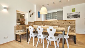 Un comedor con un banco de madera y sillas blancas, una lámpara colgante y una cocina al fondo.
