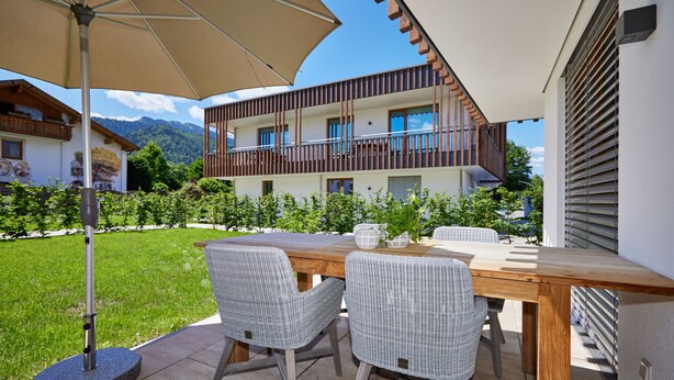 Un patio al aire libre con mesa de madera y sillas de mimbre, un paraguas grande y vistas de un edificio de dos pisos con balcones.