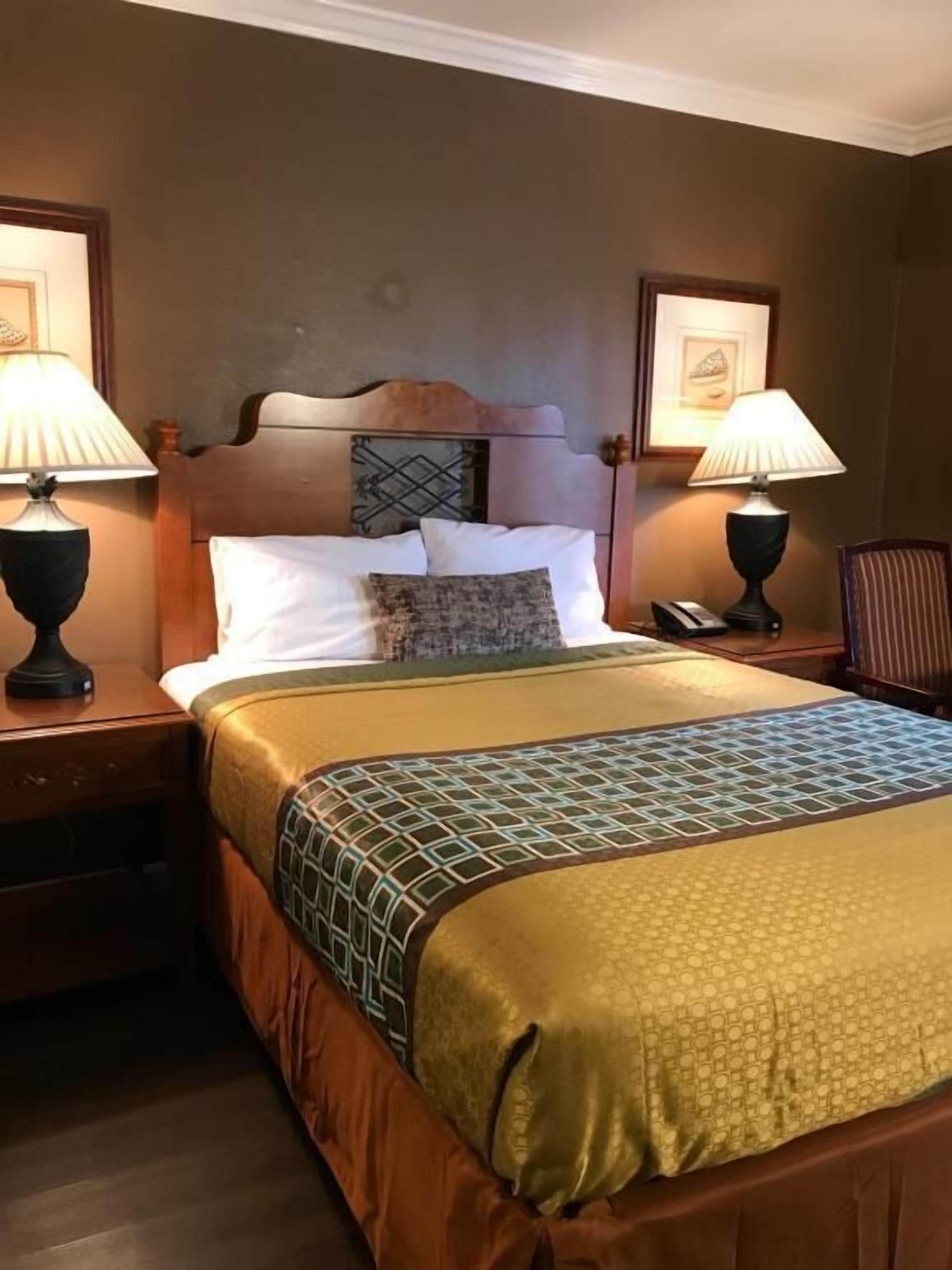 A hotel room with a large bed, two bedside tables, a nightstand, a headboard with a decorative panel, and two wall-mounted lamps.